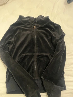 Svart velour hoodie med dragkedja - Mjuk och skön svart hoodie i velour med dragkedja framtill och fickor. Huvan har dragsko och det finns en diskret broderad logga på bröstet. Perfekt för en avslappnad och trendig look.