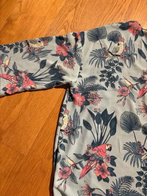 Blommig blus med papegojmotiv - Snygg blus i ljusgrått med tropiskt blommönster och papegojor i rosa, blått och vitt. Blusen har trekvartsärm och öppen front, samt dekorativ kant nertill. Perfekt till stranden i sommar att ta på över bikinin 