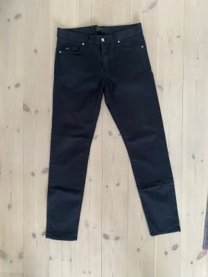 Svarta slim byxor J.Lindeberg - Svarta/ väldigt mörk grå byxor från J.Lindeberg med klassisk femficksdesign och smal passform. Byxorna har en stilren look med raka ben och är tillverkade i ett jeans liknande bomullsmaterial. Perfekta för dig som gillar minimalistisk stil och snygga detaljer. 