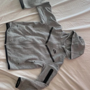 Grå huvtröja från Nike barn - Bild 3 är hur stora fickorna är. Säljer en grå huvtröja från Nike med svarta detaljer. Tröjan har dragkedja framtill och en praktisk ficka med dragkedja på ärmen. Perfekt för en sportig look eller avslappnade dagar. Barn M=137-147cm! 