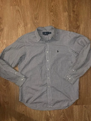 Rutig skjorta från Ralph Lauren - Klassisk rutig skjorta från Ralph Lauren i svart och vitt. Skjortan har lång ärm, knappar framtill och den ikoniska loggan broderad på bröstet. Tillverkad i bomull och har en normal passform. Perfekt för en stilren och avslappnad look. Storlek L och väldigt fint skick. Hör gärna av vid intresse!