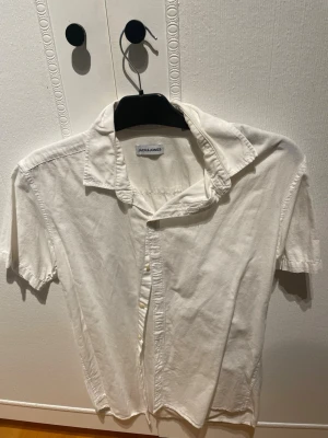 Vit kortärmad skjorta Jack & Jones - Klassisk vit skjorta från Jack & Jones i storlek M. Skjortan har korta ärmar, knappar framtill och en avslappnad krage. Tillverkad i mjuk bomull som känns skön mot huden. Perfekt för en clean och enkel stil.