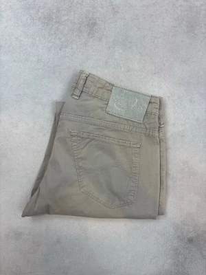 Beige chinos från Jacob Cohen - Snygga beige chinos från Jacob Cohen med raka ben och klassisk femficksdesign. Byxorna har bälteshällor, knappgylf och diskret logotyp-patch bak. Tillverkade i mjukt bomullstyg som ger en avslappnad men stilren look.