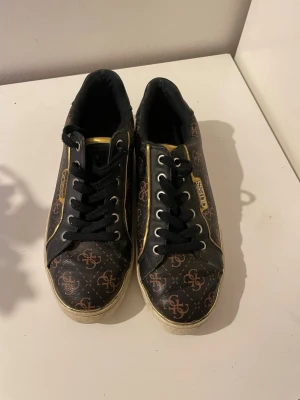 Svarta Guess sneakers med guldmönster - Snygga svarta sneakers från Guess med guldfärgade detaljer och logomönster över hela skon. Skorna har svarta snören, vita sulor och metallfärgade öglor. Materialet är konstläder med glansig finish och insidan är mjukt vadderad.