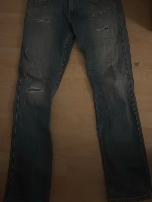 Blå slitna jeans med raka ben replay - Säljer ett par blå jeans med raka ben och slitna detaljer på både lår och knän. Jeansen har klassisk femficksmodell och en tvättad look som ger en avslappnad vibe. Perfekta för dig som gillar en trendig och casual stil.