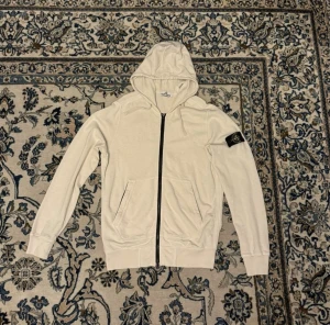 Vit hoodie med dragkedja Stone Island - Vit hoodie från Stone Island med svart dragkedja, två fickor fram och ikonisk logotyp-patch på vänster ärm. Tillverkad i mjuk bomull och har en klassisk huva. Perfekt för dig som gillar streetwear och vill ha en clean look.
