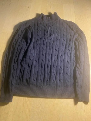 Mörkblå kabelstickad tröja - Mysig mörkblå kabelstickad tröja med halv dragkedja vid halsen och ribbade muddar. Tröjan har ett klassiskt flätmönster över hela plagget och hög krage som ger extra värme. Perfekt för kyliga dagar och enkel att matcha med jeans eller kjol.