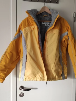 Gul vindjacka från Columbia - Snygg gul vindjacka från Columbia med gråa detaljer och dragkedja. Jackan har en mjuk grå fleece på insidan och praktiska fickor med dragkedja. Perfekt för blåsiga dagar och outdoor-äventyr. Sportig look och bekväm passform.