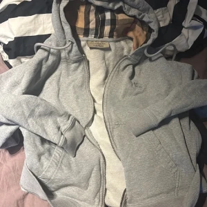 Burberry hoodie - Säljer en grå hoodie från Burberry med dragkedja framtill och klassiskt rutigt foder i huvan 