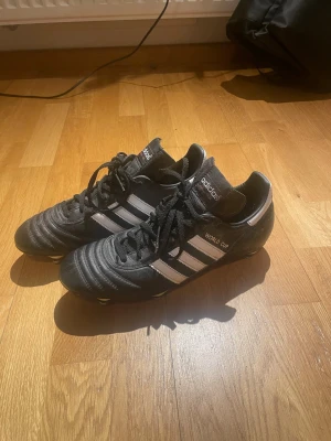 Adidas World Cup fotbollsskor copa mundial - Klassiska Adidas World Cup fotbollsskor i svart läder med vita detaljer och de ikoniska tre ränderna på sidan. Skorna har snörning, låg profil och dobbar för spel på naturgräs. Perfekta för dig som vill ha stilrena och pålitliga skor på planen. Använda tre gånger 
