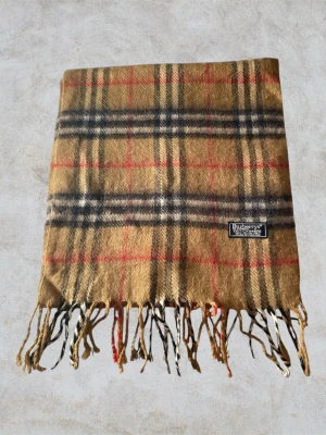 Klassisk Burberry halsduk  - Klassisk Burberry-halsduk i 100% lammull. Beige med ikoniskt rutmönster i svart, vitt och rött. Mjuk, varm och i fint skick. Perfekt för både vardag och höst-/vinterstil. Hör av er vid minsta fundering 