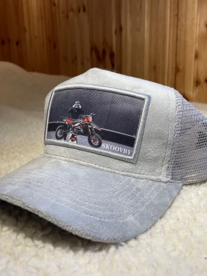 Grå truckerkeps från Skoovby - Säljer en grå truckerkeps från Skoovby med mesh på sidorna och baksidan. Framtill finns en stor patch med motiv av en motorcykel och förare. Kepsen har böjd skärm och justerbar snapback. Perfekt för dig som gillar streetwear och coola detaljer. Inte mycket andvänd! 