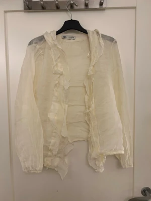 Vit volangblus från Zara - Snygg och luftig vit blus från Zara med volangdetaljer längs fram och runt kragen. Blusen är transparent och har långa ärmar med resår vid ärmslut. Perfekt att styla lager på lager för en trendig look.