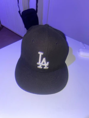 Svart LA Dodgers keps New Era - Säljer en svart keps från New Era med LA Dodgers-logga broderad i vitt framtill. Klassisk flat brim och MLB-logga baktill. Kepsen är gjord i ett tåligt tygmaterial och har en stilren look som passar till streetwear.