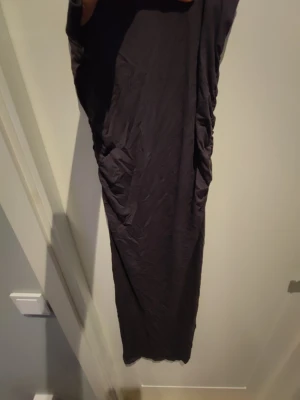 Svart lång kjol med rynkade detaljer - En svart långkjol med rynkade detaljer längs sidorna. Klänningen har en enkel och stilren design utan synliga mönster eller dekorationer. Materialet känns mjukt och stretchigt, vilket ger en bekväm passform. Jag är 166 typ och jag går lite på den om jag använder den säljer den för att den inte kommer till användning. Lite jobbig att gå i men vet inte om de är bara jag som tycker de eller inte så säljer den till ett rimligt pris köpt för 150kr typ på rea. Aldrig använd! Bara provad.