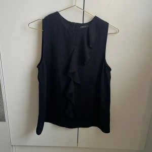 Svart ärmlös blus med volang - Snygg svart ärmlös blus från Stockh LM med volangdetalj framtill. Blusen har rund halsringning och stängs med knapp i nacken. Perfekt att styla till både jeans och kjol för en trendig look.🫰🏼💓