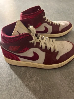 Air Jordan 1 Mid röd/vit - Snygga Air Jordan 1 Mid sneakers i rött och vitt med klassisk Nike Swoosh på sidan. Skorna har vita snören, perforerad tåbox och detaljer i röd mocka och läder. Tungan är beige med Jumpman-logga och yttersulan är gulaktig. Perfekta för dig som gillar streetwear.