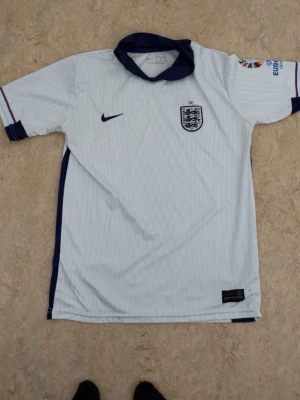 England Nike vit fotbollströja - Snygg vit England fotbollströja från Nike med blå detaljer och broderat landslagsmärke på bröstet. Tröjan har korta ärmar och sportig passform, perfekt för match eller träning. Materialet är lätt och andas bra. Vid mer info kontakta gärna! ABS! Pris går att diskutera! 