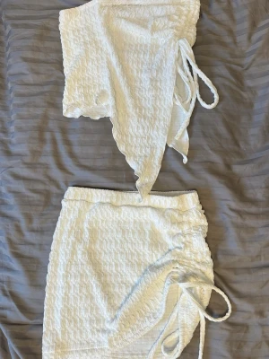 Vitt tvådelat set med struktur - Säljer ett vitt tvådelat set med asymmetrisk topp och kjol i stretchigt, strukturerat tyg. Toppen är axelbandslös och har snörning i sidan, medan kjolen har matchande snörningsdetalj. Perfekt för dig som gillar trendiga och unika plagg.