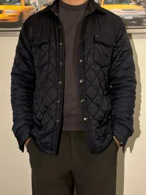 Marinblå quiltad  Tommy Hilfiger - Snygg marinblå quiltad från Tommy Hilfiger med tryckknappar framtill och bröstficka med liten logga. Jackan har krage och ett rutmönster som ger en modern look. Perfekt för dig som vill ha en lätt och stilren jacka till vardags.