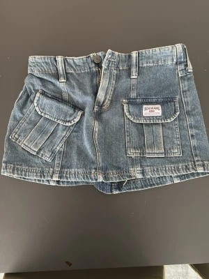 Blå jeanskjol från Urban Outfitters BDG - Säljer en blå jeanskjol från BDG med två stora fickor framtill och bälteshällor. Kjolen har knapp och dragkedja i midjan samt en avslappnad passform. Perfekt till sommaren för en cool och avslappnad look.