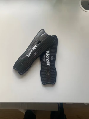 Monolit benskydd i kolfiber - Säljer ett par svarta benskydd från Monolit, tillverkade i kolfiber med en snygg, sportig design. Lätta och smidiga för bästa rörelsefrihet, perfekta för fotboll eller andra sporter där skydd behövs. Snyggt Monolit-tryck på framsidan.