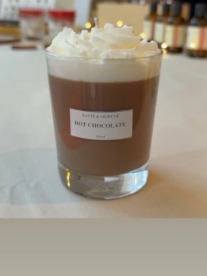 Doftljus - hot chocolate 🤎 - Säljer ett doftljus i glas som ser ut som varm choklad med vispgrädde. Ljuset är brunt med vit topp och etikett på framsidan där det står 'Hot Chocolate'. Perfekt för dig som vill ha mysig chokladdoft hemma.