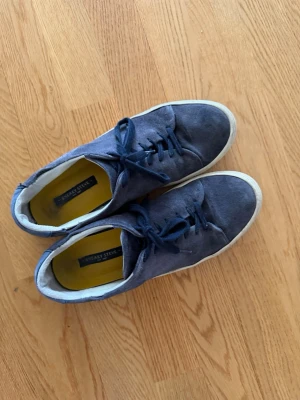 Blå mockasneakers från Sneaky Steve - Mörkblå sneakers i mocka från Sneaky Steve med vita sulor och matchande blå skosnören. Låg modell med gul insida och diskret logga på innersulan. Perfekta för dig som gillar stilrena och klassiska sneakers med en twist.