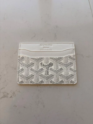 Vit kortfodral 1:1 Go*ard - Snyggt vitt kortfodral med klassiskt mönster i svart och grått. Plånboken har flera kortfack och är tillverkad i exklusivt läder. Perfekt för dig som vill ha en stilren och lyxig accessoar i fickan.