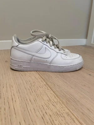 Nike Air Force 1 vita sneakers - Klassiska vita Nike Air Force 1 sneakers i lågt utförande. Skorna har en stilren design med perforerad tå, vita skosnören och den ikoniska swoosh-loggan på sidan. Tillverkade i läder med en robust gummisula för extra komfort.