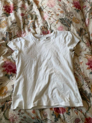 Vit basic t-shirt från H&M - En enkel vit t-shirt från Kappahl med rund hals och korta ärmar. Tillverkad i mjuk bomull och har en klassisk passform som funkar till det mesta. Perfekt basplagg för garderoben.