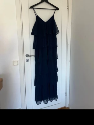Marinblå långklänning med volanger - Säljer en marinblå långklänning med tunna axelband och flera lager volanger. Klänningen har en luftig och flowy känsla, perfekt för sommarens alla tillfällen. Materialet är lätt och ger ett elegant fall.
