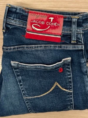 Jacob Cohën mörkblå jeans - Snygga mörkblå jeans från Jacob Cohën med klassisk femficksdesign. Jeansen har en röd patch med logga bak och gul brodyr på bakfickan. Tillverkade i denim med kontrastsömmar och detaljer som sticker ut.