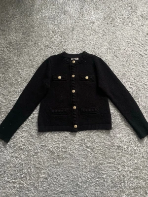 Svart kofta med guldknappar H&M - Stilren svart kofta från H&M med dekorativa guldknappar framtill och på fickorna. Koftan har rund halsringning, två bröstfickor och två fickor nedtill. Materialet är stickat och ger en klassisk look som passar till många outfits. 