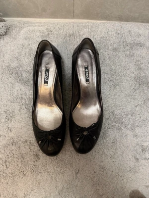 Svarta ballerinaskor från Ecco - Stilrena svarta ballerinaskor från Ecco med rund tå och dekorativ rosett framtill. Skorna är i läder och har en klassisk, tidlös design som passar till många olika outfits. Perfekta för dig som gillar enkel och snygg stil.