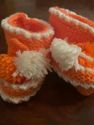 Stickade orangea tofflor med tofs - Säljer ett par ny  handstickade tofflor i orange och vitt med fluffiga tofsar framtill. De har en söt, vågig kant och är gjorda i mjukt garn som känns skönt mot huden. Perfekta för att hålla fötterna varma och mysiga hemma.