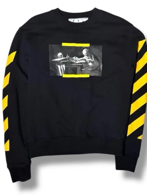 Svart Off-White hoodie med konsttryck - Svart hoodie från Off-White med konstnärligt tryck av två skelett på bröstet och gula detaljer. Ärmarna har Off-Whites ikoniska gula diagonala ränder. Rund halsringning och ribbade muddar. Perfekt för dig som gillar streetwear och unika plagg.