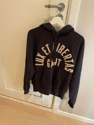 Svart hoodie från GANT med tryck - Snygg svart hoodie från GANT med texten 'LUX ET LIBERTAS' i vitt tryck över bröstet. Klassisk huva med dragsko och stor magficka framtill. Perfekt för en avslappnad och cool stil. Tillverkad i mjukt bomullsmaterial.
