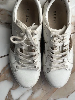 Vita Guess sneakers med logga - Snygga vita sneakers från Guess med klassisk låg profil och bruna paneler på sidorna med Guess-logga. Skorna har vita snören och detaljer i syntetläder. Perfekta för en trendig och avslappnad stil.
