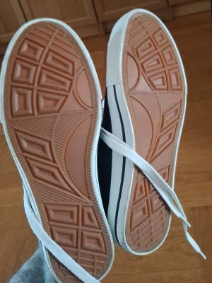 Svarta canvas sneakers med vita detaljer - Snygga svarta sneakers i klassisk modell med vita snören och sula. Skorna har en vit rand längs sulan och ovandel i canvas. Yttersulan är i gummi med mönstrad undersida för bra grepp. Perfekta för en avslappnad streetstil.