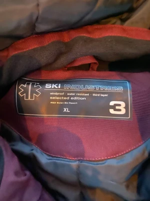 Vinröd skidjacka från Ski Industries - Säljer en snygg vinröd skidjacka från Ski Industries i storlek XL. Jackan har rosa paneler på sidorna och ärmarna, huva och dragkedja framtill. Materialet är vindtätt och vattenavvisande, perfekt för blåsiga dagar. Märkt med 'selected edition'.helt ny 