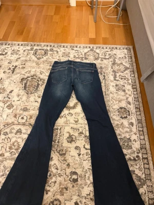 Mörkblå bootcut jeans med slitningar - Säljer ett par mörkblå bootcut jeans med snygga slitningar och dubbla knappar i midjan. Jeansen har klassiska fem fickor och markerade sömmar. Perfekta för dig som gillar en retroinspirerad look och vill ha ett par jeans som sticker ut.