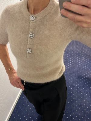 Ny Beige stickad topp tröja  med smyckesknappar hm Stl s - Säljer en fluffig, beige  stickad topp från  HM kort ärmar och rund hals. Toppen har tre dekorativa smyckesknappar framtill som ger en lyxig touch. Materialet ser ut att vara en mjuk ull- eller mohairblandning.