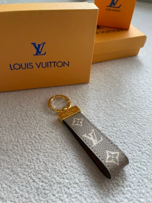 Vuitton nyckelring monogram - Snygg nyckelring från Vuitton med klassiskt monogrammönster i beige och vitt på canvas. Detaljer i guldfärgad metall och präglad logotyp på metallplattan. Perfekt accessoar för att ge din nyckelknippa en lyxig touch.