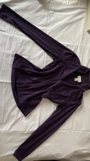 Lila träningsjacka från H&M - Snygg lila träningsjacka från H&M med dragkedja framtill och långa ärmar. Jackan har en figurnära passform och är tillverkad i ett stretchigt material som passar perfekt för träning eller sportaktiviteter.
