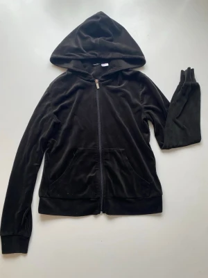 Svart velourhoodie med dragkedja - Mjuk och skön svart hoodie i velour med dragkedja framtill och två fickor. Klassisk huva och långa ärmar. Perfekt för en avslappnad och trendig look. Passar till både jeans och mjukisbyxor.