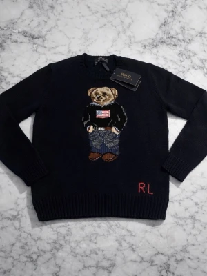 Mörkblå stickad tröja Polo Bear RL - Mörkblå stickad tröja från Polo Ralph Lauren med ikoniska Polo Bear-motivet framtill och röda RL-bokstäver nedtill. Tröjan har rund halsringning och ribbade muddar. Perfekt för dig som gillar klassisk och lekfull stil.