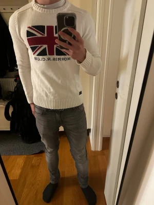 Vit stickad tröja Morris R Club - Vit stickad tröja från Morris R Club med Union Jack-flagga i rött, svart och vitt på bröstet | Rund halsringning | Ribbstickade muddar vid ärmslut och nederkant | Klassisk och stilren design | Mått: midja 46 cm | underarm 48 cm | längd 63 cm | 