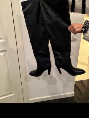 Svarta overknee stövlar med klack - Säljer ett par svarta overknee stövlar i slätt konstläder med spetsig tå och hög blockklack. Stövlarna har en stilren design och sitter snyggt över knät. Perfekta för att lyfta vilken outfit som helst.