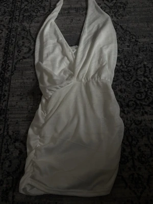 Vit strandklänning med djup urringning - Säljer en vit strandklänning i mesh-liknande material med djup urringning och halterneck. Klänningen har rynkade detaljer på sidan och är lätt transparent, perfekt att dra över bikinin på stranden.